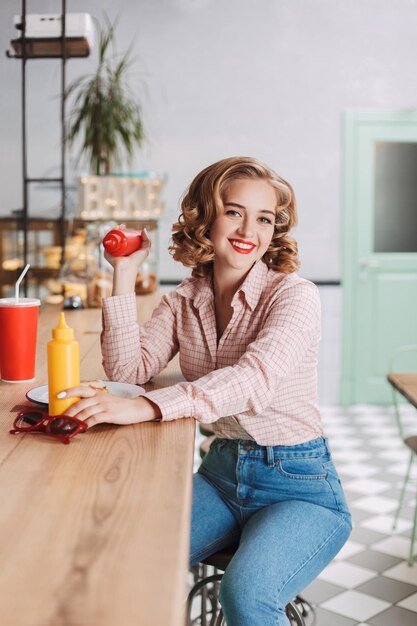 Jonge lachende dame in shirt en spijkerbroek zit aan de bar met ketchup en mosterdflessen en kijkt graag in de camera in café