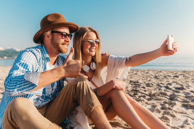 Jonge lachende gelukkig man en vrouw in zonnebril zittend op zand strand selfie foto op telefooncamera te nemen