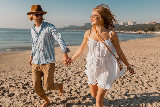 Jonge lachende gelukkig man in hoed en blonde vrouw in witte jurk samen uitgevoerd op strand op zomervakantie reizen