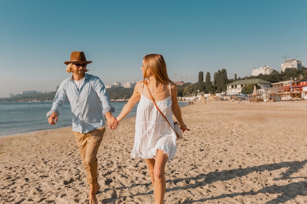 Jonge lachende gelukkig man in hoed en blonde vrouw loopt samen op het strand op zomervakantie reizen