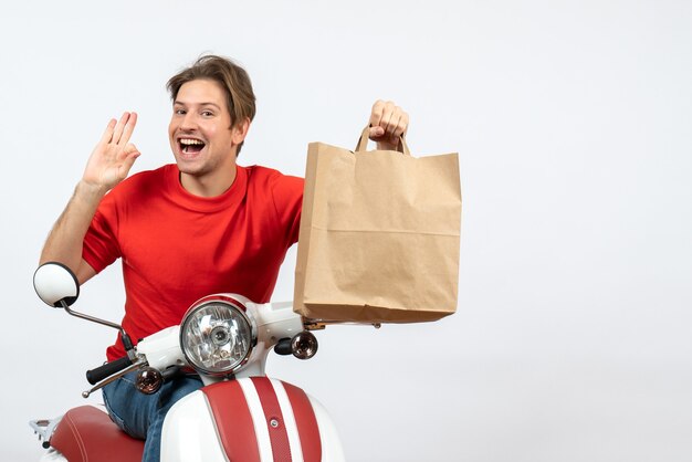 Jonge lachende koerier man in rood uniform zittend op scooter houden papieren zak en bril gebaar maken op gele muur