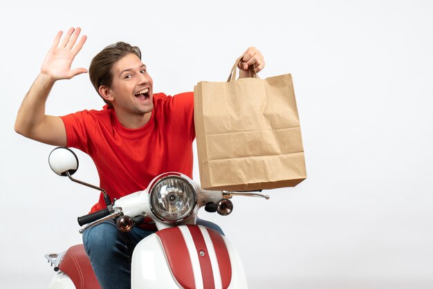 Jonge lachende koerier man in rood uniform zittend op scooter met papieren zak hallo zeggen op witte muur