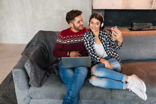 Jonge lachende man en vrouw om thuis te zitten in de winter, laptop houden, luisteren naar koptelefoon, koppel op vrije tijd samen, selfie foto maken op smartphone camera, gelukkig, positief, dating