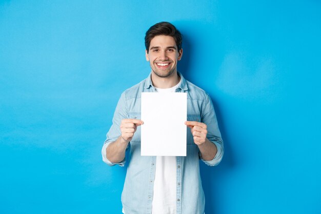 Jonge lachende man in casual outfit, met blanco vel papier met uw advertentie, staande over blauwe achtergrond.