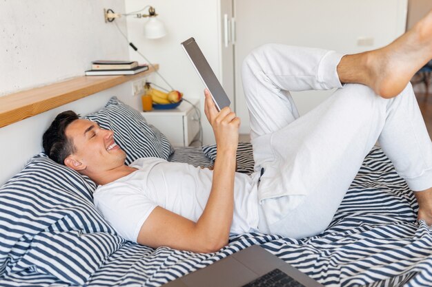 Jonge lachende man in casual pyjama outfit zittend in bed in de ochtend met tablet, freelancer thuis