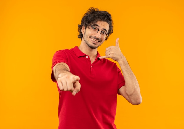 Jonge lachende man in rood shirt met optische bril gebaren bel me en punten geïsoleerd op oranje muur