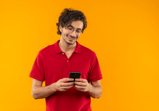 Jonge lachende man in rood shirt met optische bril houdt en kijkt naar telefoon geïsoleerd op oranje muur