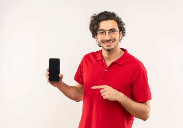 Jonge lachende man in rood shirt met optische bril houdt en wijst op telefoon geïsoleerd op een witte muur