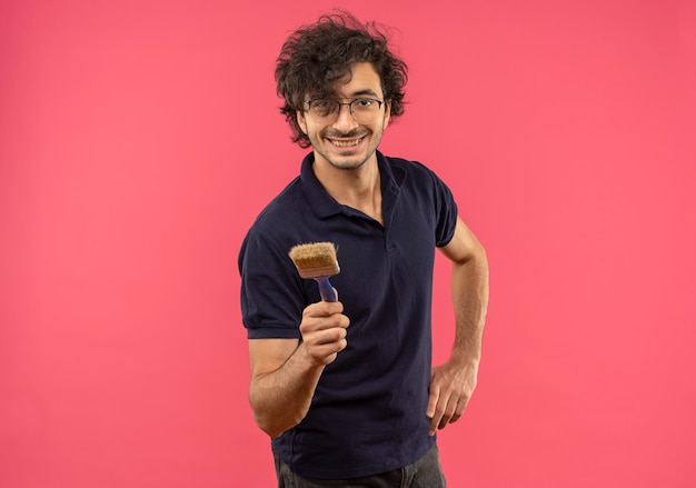 Jonge lachende man in zwart shirt met optische bril houdt borstel en legt hand op taille geïsoleerd op roze muur