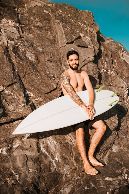 Jonge lachende man met surfplank in de buurt van stenen