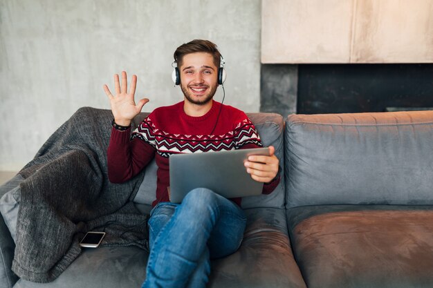 Jonge lachende man zit thuis in de winter, zwaaiende hand, hallo zeggen, rode trui dragen, bezig met laptop, freelancer, koptelefoon luisteren, student online studeren, in de camera kijken