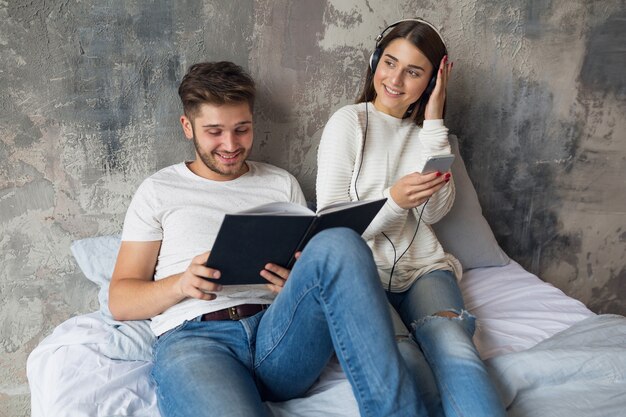 Jonge lachende paar zittend op bed thuis in casual outfit leesboek dragen jeans, man leesboek, vrouw luisteren naar muziek op koptelefoon, romantische tijd samen doorbrengen