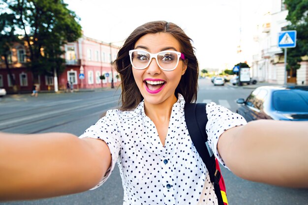 Jonge lachende tiener gelukkige vrouw selfie maken op straat, leng haren, lichte make-up en schattige heldere bril, alleen reizen, plezier, positieve stemming, vreugde, vakantie