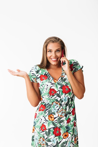 Jonge lachende vrouw in kleurrijke jurk die gelukkig in de camera kijkt terwijl ze op mobiele telefoon praat over witte achtergrond
