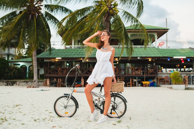 Jonge lachende vrouw in witte jurk rijden op tropisch strand op fiets zonnebril reizen op zomervakantie in Thailand