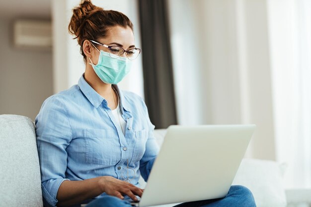 Jonge lachende vrouw met gezichtsmasker met behulp van computer terwijl u ontspant in de woonkamer