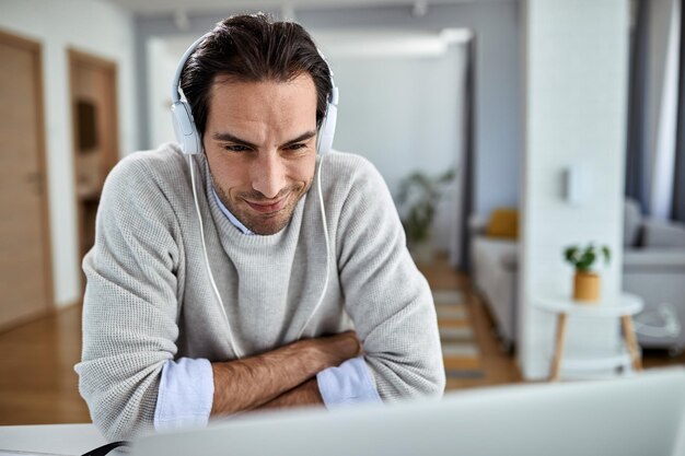 Jonge lachende zakenman met koptelefoon die iets op een computer leest terwijl hij thuis werkt