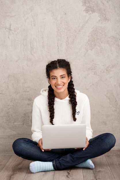 Gratis foto jonge laptop van de vrouwenholding, die op vloer over beige muur zit