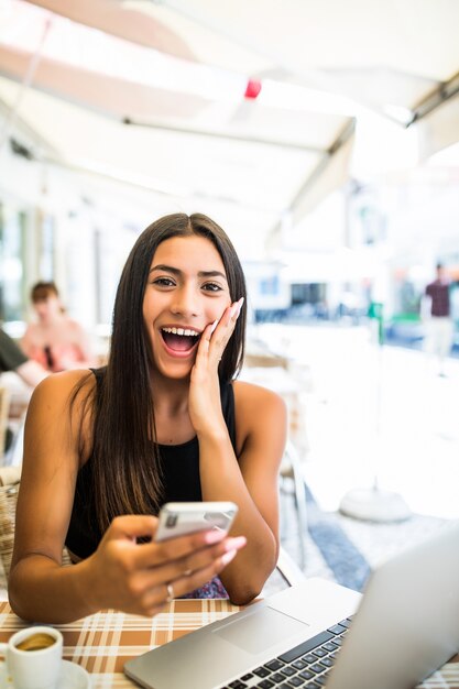 Jonge Latijns-vrouw krijgt geschokt door nieuws in haar telefoon buiten in café. Geschokt gezicht van een meisje dat op een terras zit met haar telefoon in handen.