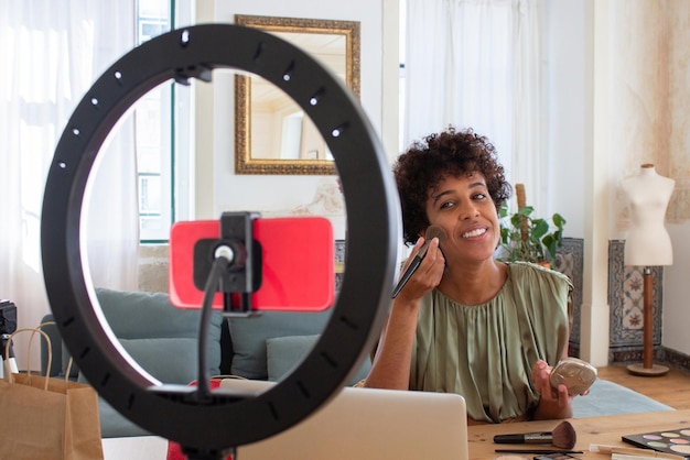 Jonge Latijnse vrouw die poeder op haar gezicht aanbrengt voor beautyblog. Glimlachende vrouw zittend aan tafel in een gezellige kamer met poederdoos en borstel kijkend naar telefooncamera die video opneemt. Make-up en cosmetica b