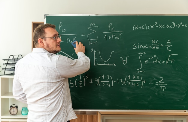 Jonge leraar met een bril die zich achter het zicht voor het bord in de klas bevindt en naar de zijkant kijkt die het schoolbord schoonmaakt met krijtwisser