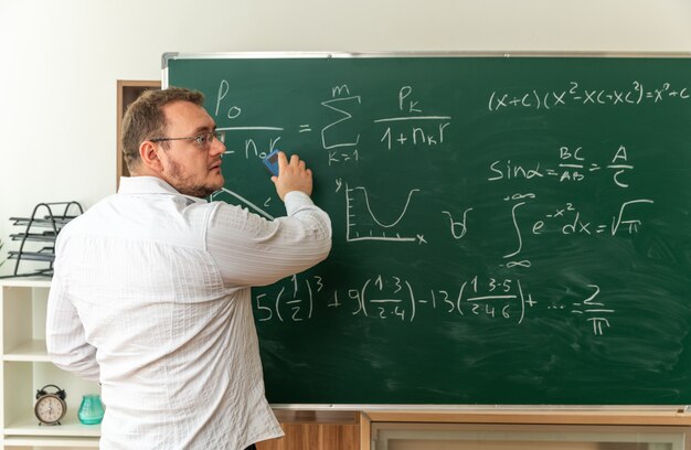 jonge leraar met een bril die zich achter het zicht voor het bord in de klas bevindt en naar de zijkant kijkt die het schoolbord schoonmaakt met krijtwisser