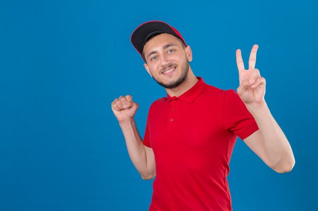 Jonge levering man dragen rode polo shirt en pet camera kijken met glimlach op gezicht verhogen vuist en overwinning teken winnaar concept tonen over geïsoleerde blauwe achtergrond