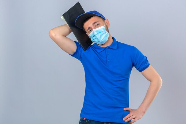 Jonge levering man met blauw poloshirt en pet in beschermend medisch masker staan met klembord op schouder moe en verveeld kijken over geïsoleerde witte achtergrond