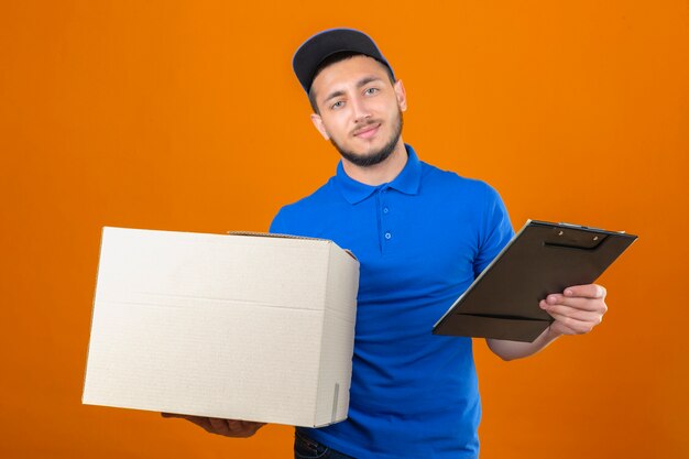 Jonge levering man met blauw poloshirt en pet permanent met kartonnen doos en klembord camera kijken met glimlach op gezicht over geïsoleerde oranje achtergrond