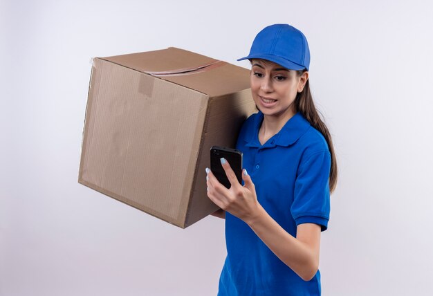 Jonge levering meisje in blauw uniform en pet bedrijf grote kartonnen doos kijken scherm van mobiele telefoon bezorgd en verward