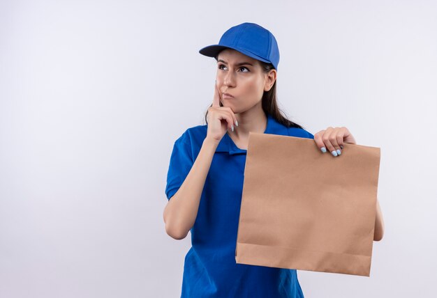 Jonge levering meisje in blauw uniform en pet houden papier pakket opzij kijken met peinzende uitdrukking op gezicht, denken