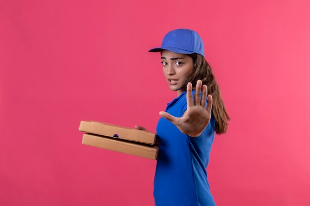 Jonge levering meisje in blauw uniform en pet houden van pizzadozen permanent met open hand doen stopbord met ernstige en zelfverzekerde expressie verdediging gebaar op roze achtergrond