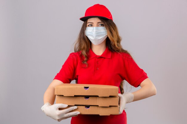 Jonge levering vrouw met krullend haar dragen rode polo shirt en pet in medische beschermend masker en handschoenen staan met pizzadozen camera kijken met glimlach op gezicht over geïsoleerde witte CHTERGRO