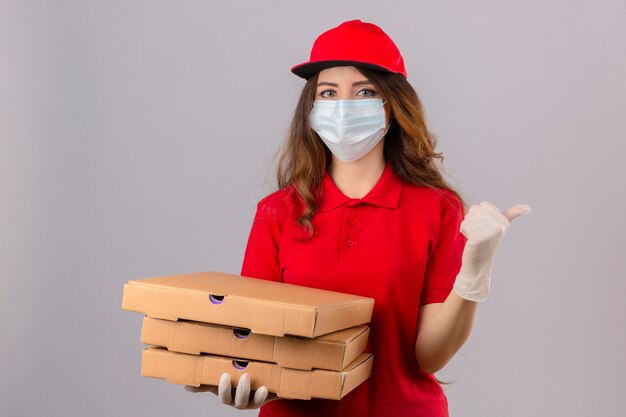 Jonge levering vrouw met krullend haar dragen rode poloshirt en pet in medische beschermend masker en handschoenen staan met pizzadozen wijzen en tonen met duim omhoog aan de kant met blij gezicht smi