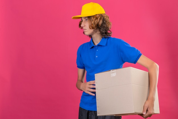 Gratis foto jonge leveringsmens die blauw poloshirt en gele pet draagt die grote kartondoos in handen houden die met ernstig gezicht opzij kijken dat zich over geïsoleerde roze achtergrond bevindt