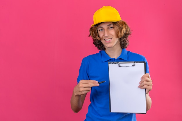 Jonge leveringsmens die blauw poloshirt en gele pet draagt die Klembord en pen in handen houdt die om handtekening vraagt die camera bekijkt die vriendelijk over geïsoleerde roze achtergrond glimlacht