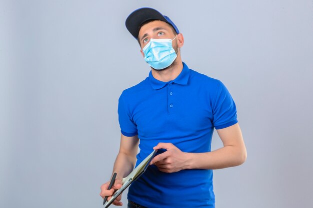 Jonge leveringsmens die blauw poloshirt en pet in beschermend medisch masker dragen die zich met klembord bevinden die omhoog denken verbaasd over geïsoleerde witte achtergrond kijken