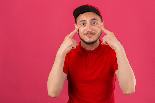 Jonge leveringsmens die rood poloshirt en pet draagt die zijn ogen richten die camera over geïsoleerde roze achtergrond glimlachen bekijken