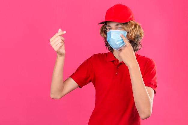 Jonge leveringsmens die rood poloshirt en pet in medisch masker draagt die een geldgebaar doet die zelfverzekerd over geïsoleerde roze achtergrond kijkt