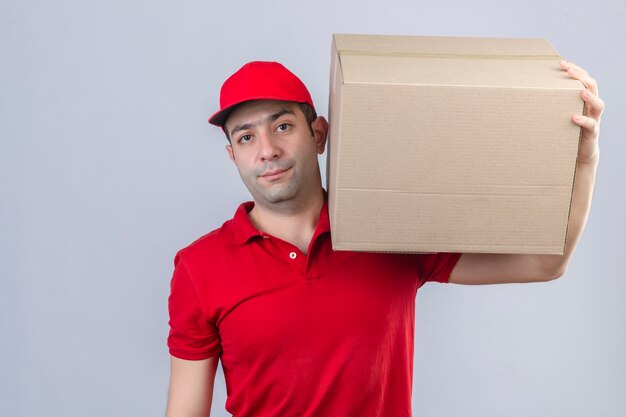 Jonge leveringsmens in rood poloshirt en GLB-de dozen van het holdingskarton met ernstig gezicht die zich over geïsoleerde witte muur bevinden