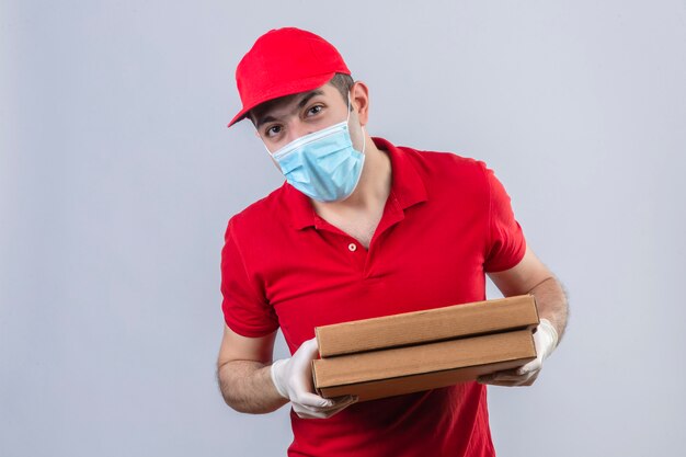 Jonge leveringsmens in rood poloshirt en GLB in medische de pizzadozen die van de maskerholding onderzoekend de camera bekijken die zich over geïsoleerde witte muur bevinden