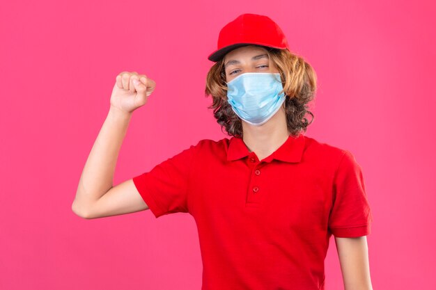 Jonge leveringsmens in rood uniform die medisch masker draagt dat vuist trots en zelfverzekerd winnaarconcept opheft dat zich over geïsoleerde roze achtergrond bevindt