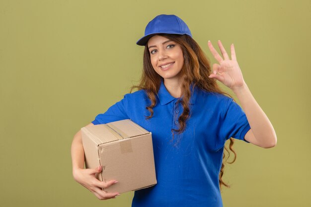 Jonge leveringsvrouw die blauw poloshirt en GLB dragen die zich met kartondoos bevinden die ok teken doen glimlachen vriendelijk over geïsoleerde groene achtergrond