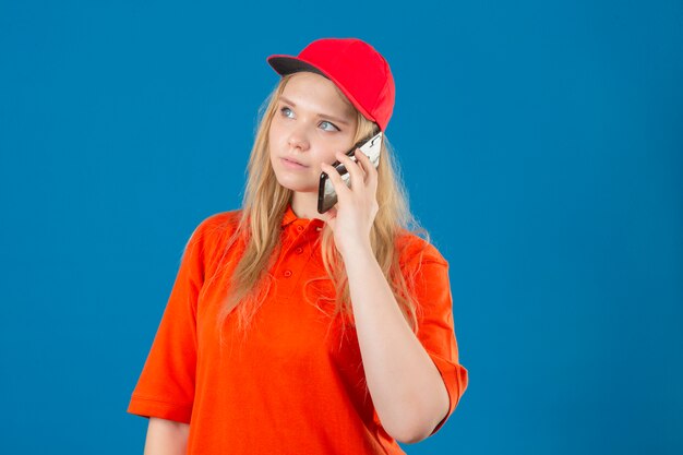 Jonge leveringsvrouw die oranje poloshirt en een rode pet draagt die op mobiele telefoon met zelfverzekerde blik over geïsoleerde blauwe achtergrond spreekt