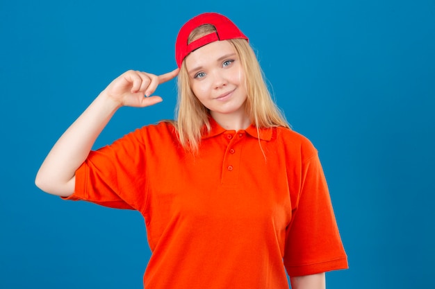 Jonge leveringsvrouw die oranje poloshirt en een rode pet draagt die tempel met vinger het denken richt dat zich op een taak over geïsoleerde blauwe achtergrond concentreert
