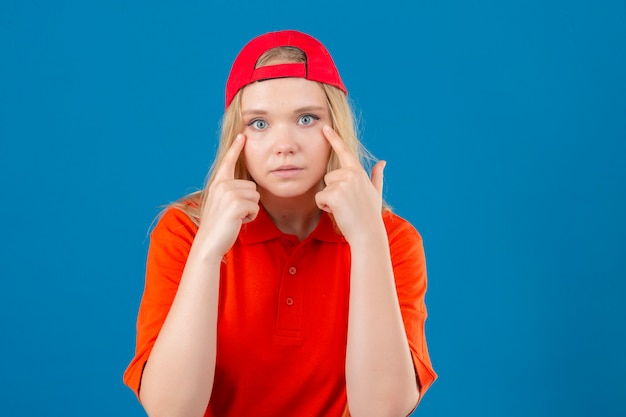 Gratis foto jonge leveringsvrouw die oranje poloshirt en een rood glb draagt die camera bekijkt die haar ogen over geïsoleerde blauwe achtergrond richt