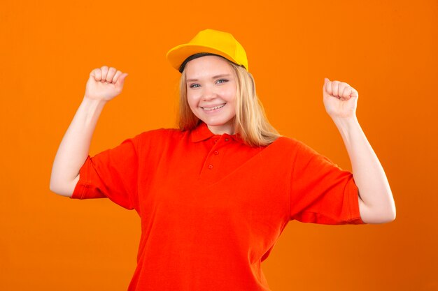 Jonge leveringsvrouw die rood poloshirt en geel GLB draagt dat vuisten opheft na een de winnaarconcept van het overwinnings blij gezicht over geïsoleerde witte achtergrond
