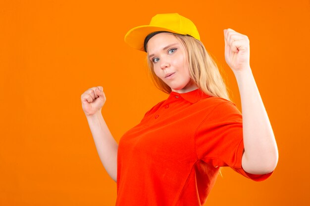 Jonge leveringsvrouw die rood poloshirt en geel GLB dragen die vriendschappelijke status met opgeheven vuistenwinnaarconcept glimlachen over geïsoleerde oranje achtergrond