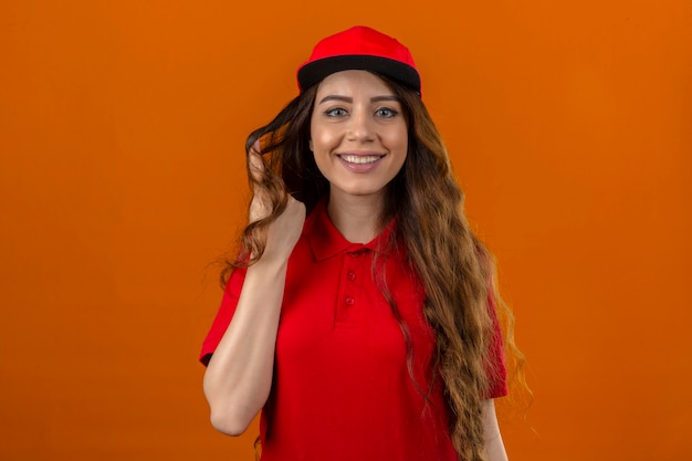 Jonge leveringsvrouw die rood poloshirt en GLB draagt dat een haarlok op de vinger verdraait die vrolijk over geïsoleerde oranje achtergrond glimlacht