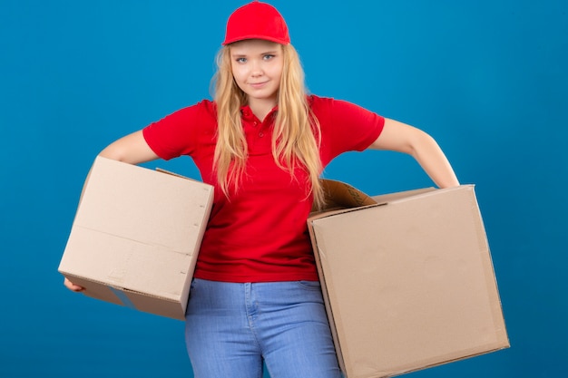 Gratis foto jonge leveringsvrouw die rood poloshirt en glb draagt die zich met grote kartondozen bevinden die camera met glimlach op gezicht over geïsoleerde blauwe achtergrond bekijken